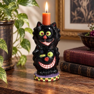 May include: A ceramic candle holder shaped like two stacked black cats with wide grins, bright green eyes, and red noses. The cats feature textured black fur, and the base is decorated with a colorful checkered pattern. An orange candle is lit on top.