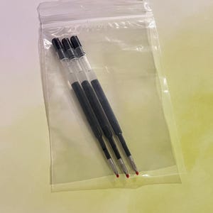 May include: Three black ink pen refills with clear plastic barrels and small red protective caps on the tips, enclosed within a transparent, resealable plastic bag. The refills feature a slim, cylindrical design with black ink visible inside.