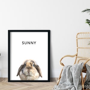 Op de afbeelding: Een zwart ingelijst print met een witte achtergrond en een cartoon afbeelding van een bruin en wit konijn met de naam "SUNNY" in zwart bovenaan.