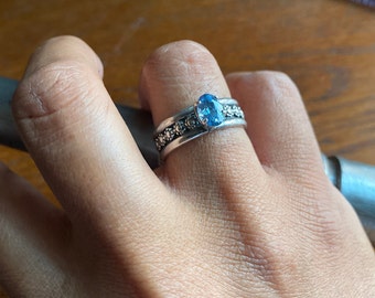 Puede incluir: Un anillo de plata con una piedra ovalada azul engastada en el centro. El anillo tiene una banda decorativa con pequeñas cuentas.