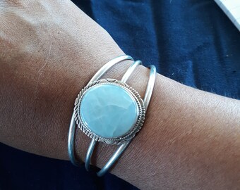 May include: A silver bracelet with a round, light blue stone set in the center. The bracelet is made of three bands of silver that are intertwined.