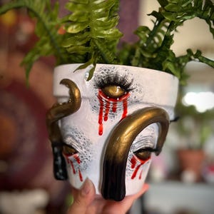 May include: White ceramic planter with a face design, featuring gold accents and red tear streaks. The planter holds green fern plants. The face has detailed eyes with long eyelashes. A unique and artistic decorative item.