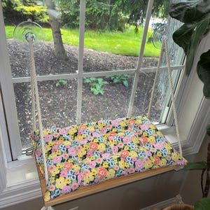 May include: A wooden window perch with a floral cushion. The cushion features a pattern of pink, yellow, and blue flowers. The perch is suspended by white rope and attached to the window with clear suction cups. The window overlooks a green yard.