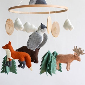 May include: A wooden baby mobile with felt woodland animals, including a fox, owl, deer, and bear. The mobile also features felt clouds, trees, and a mountain.