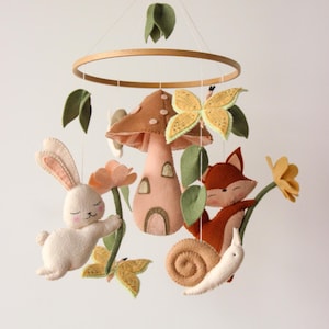 May include: A whimsical baby mobile featuring felt woodland creatures. The mobile includes a white bunny, a fox, a snail, a mushroom house, and a butterfly, all suspended from a wooden hoop with green leaves and flowers.