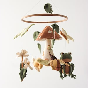May include: A whimsical felt mobile featuring a mushroom house, a frog, bees, butterflies, and flowers. The mobile is suspended from a wooden ring and includes green leaves and delicate white flowers. The color palette includes shades of brown, green, and cream.