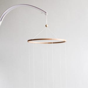 Può includere: Mobile per bambini con un anello di legno e fili sospesi. Il mobile è sospeso a un braccio curvo bianco. L'anello è in legno marrone chiaro e ha diversi fili bianchi.