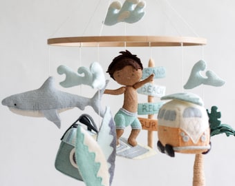 May include: A felt baby mobile with a surfer boy, a shark, a surfboard, a van, a palm tree, and clouds. The mobile is made of felt and has a wooden ring.