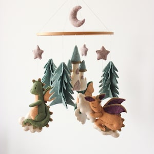 May include: A whimsical baby mobile featuring felt dragons, trees, a castle, stars, and a crescent moon. The dragons are in shades of green, orange, and purple. The trees are teal, and the castle is cream-colored. The mobile hangs from a wooden ring.