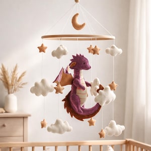May include: A baby mobile featuring a purple felt dragon, brown wings, and a crescent moon. The mobile includes felt stars and clouds, suspended from a wooden ring. The mobile hangs above a wooden crib.