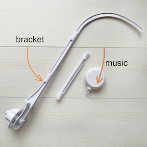 May include: A disassembled white baby mobile arm and music box. The mobile arm is curved and has a bracket. The music box is round. The components are on a light wood-grain surface.