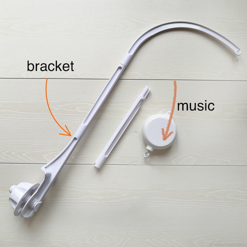 May include: A disassembled white baby mobile arm and music box. The mobile arm is curved and has a bracket. The music box is round. The components are on a light wood-grain surface.