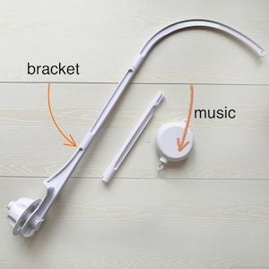 May include: A disassembled white baby mobile with the word "bracket" and "music" in the image. The mobile includes a curved arm, a central support, and a music box component. The components are arranged on a light wood-grain surface.