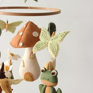 May include: A whimsical baby mobile featuring felted woodland creatures. The mobile includes a brown mushroom house, green frog, butterflies, and leaves, all suspended from a wooden hoop. The color palette is soft and natural.
