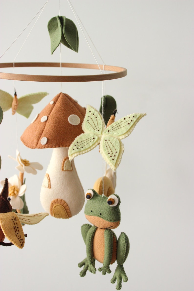 May include: A whimsical baby mobile featuring felted woodland creatures. The mobile includes a brown mushroom house, green frog, butterflies, and leaves, all suspended from a wooden hoop. The color palette is soft and natural.