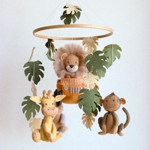 May include: A wooden baby mobile with felt animal toys. The mobile features a lion in a jeep, a giraffe, and a monkey. The animals are surrounded by green and beige felt leaves.