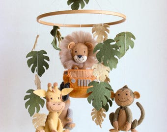 May include: A wooden baby mobile with felt animal toys. The mobile features a lion in a jeep, a giraffe, and a monkey. The animals are surrounded by green and beige felt leaves.