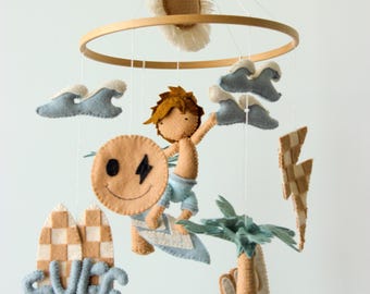 May include: A handcrafted baby mobile featuring a surfer riding a wave, a sun, clouds, and surfboards. The mobile is made of felt in shades of tan, blue, and white. The word "SURF" is spelled out on the surfboard.