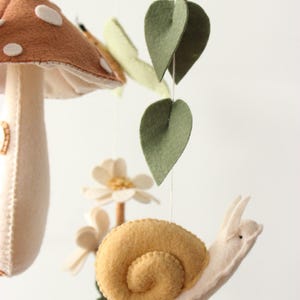 May include: A whimsical felt mobile featuring a mushroom with white spots, a snail, leaves, and flowers. The mobile is suspended from a wooden ring. The color palette includes brown, beige, yellow, and green.