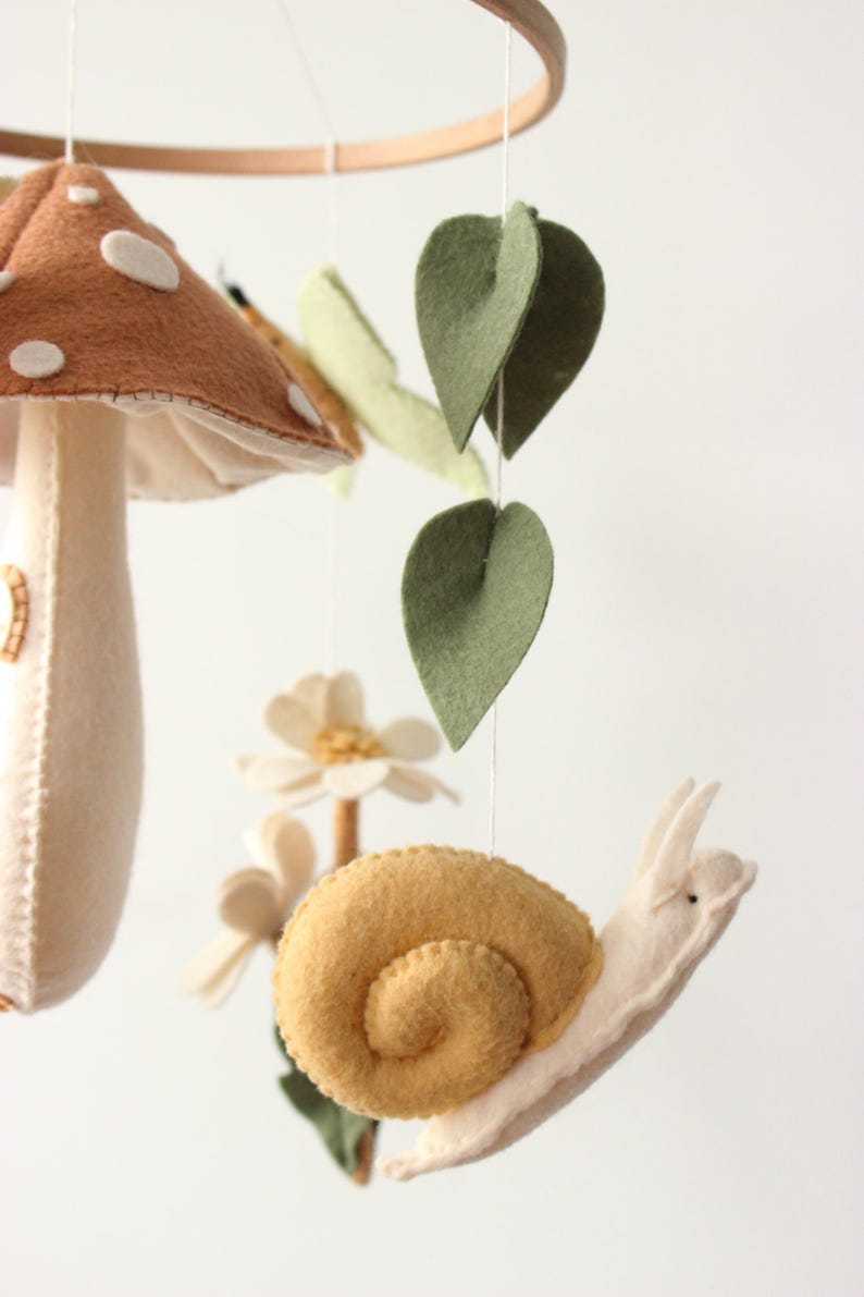 May include: A whimsical felt mobile featuring a mushroom with white spots, a snail, leaves, and flowers. The mobile is suspended from a wooden ring. The color palette includes brown, beige, yellow, and green.