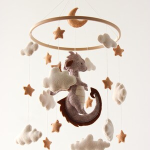 May include: A baby mobile featuring a felt dragon, clouds, stars, and a crescent moon. The dragon is gray with brown accents, and the stars and moon are tan. The mobile hangs from a wooden ring, creating a whimsical nursery decoration.