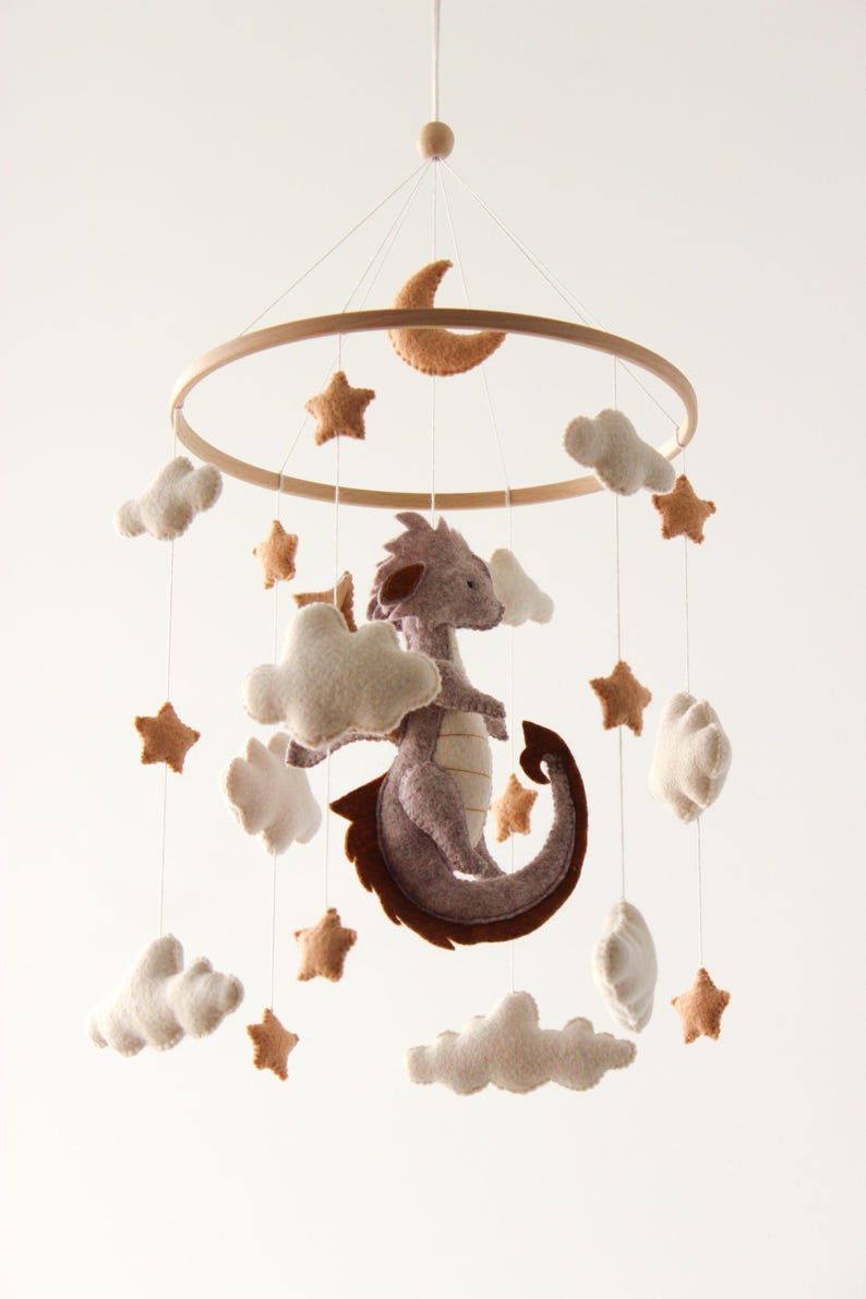 May include: A baby mobile featuring a felt dragon, clouds, stars, and a crescent moon. The dragon is gray with brown accents, and the stars and moon are tan. The mobile hangs from a wooden ring, creating a whimsical nursery decoration.