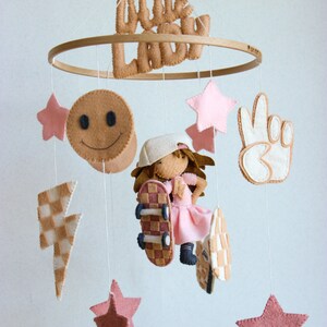 Puede incluir: Un móvil de bebé de madera con una muñeca de fieltro de una niña que monta en patineta, una cara sonriente, un rayo, un signo de paz y cinco estrellas rosas. El móvil está etiquetado como "Little Lady".