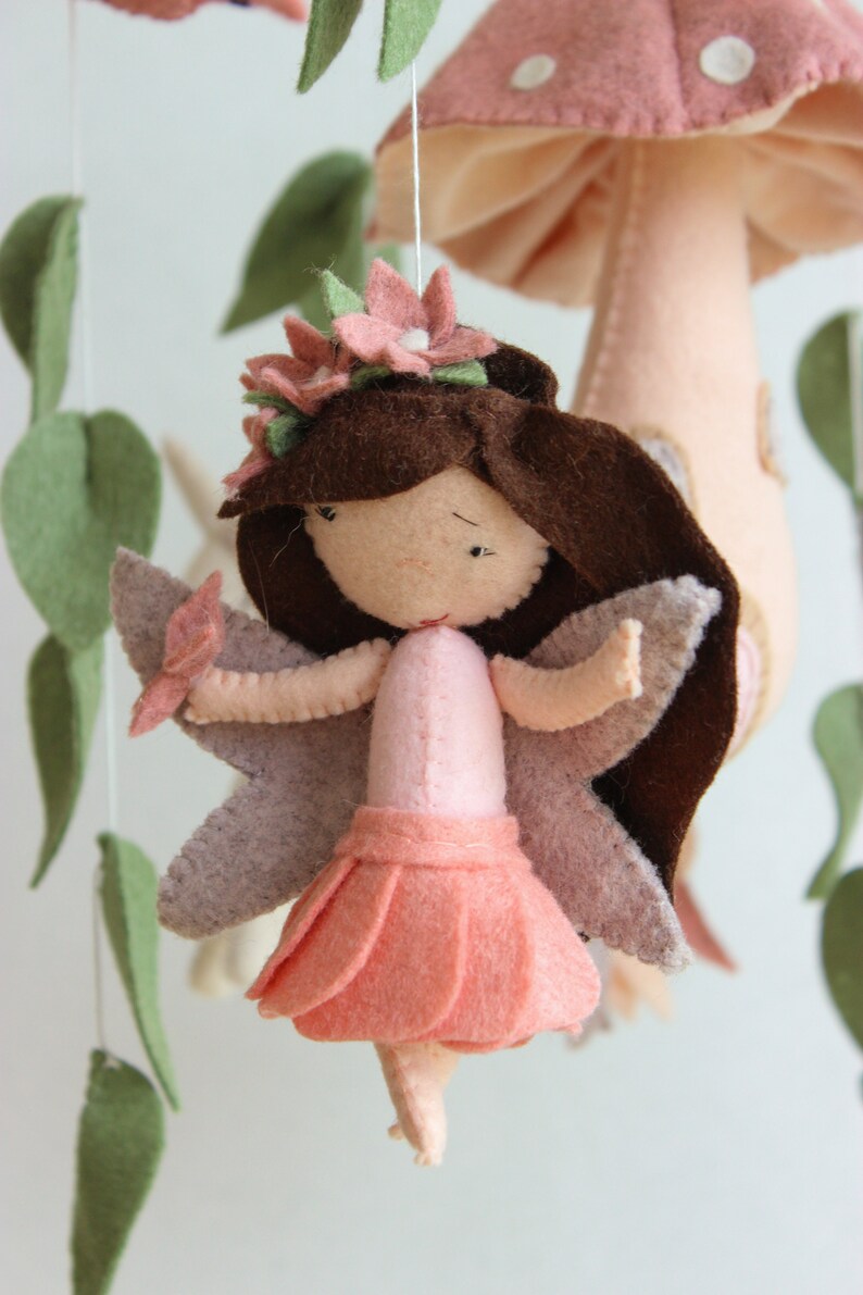 May include: A felt fairy mobile featuring a pink mushroom, green leaves, and a pink fairy with brown hair and pink wings.
