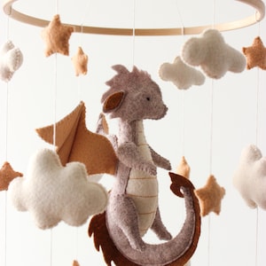 May include: A handmade baby mobile featuring a felt dragon with brown wings and tail, surrounded by felt stars, clouds, and a crescent moon. The mobile is suspended from a wooden hoop, creating a whimsical nursery decoration.