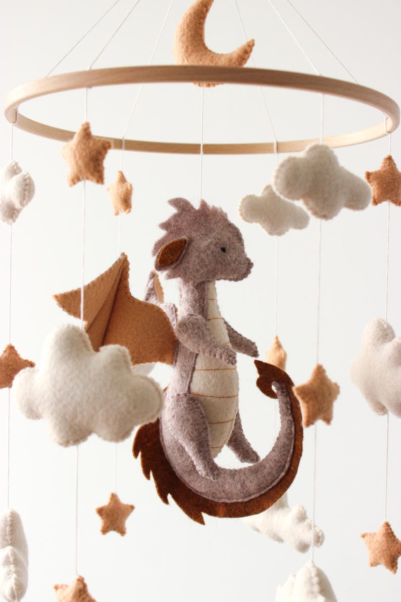 May include: A handmade baby mobile featuring a felt dragon with brown wings and tail, surrounded by felt stars, clouds, and a crescent moon. The mobile is suspended from a wooden hoop, creating a whimsical nursery decoration.