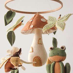 May include: A whimsical baby mobile featuring felted woodland creatures and a mushroom house. The mobile includes a brown mushroom house with white spots, a green frog, a butterfly, and a beetle, all suspended from a wooden ring.