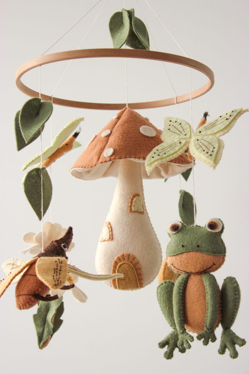 May include: A whimsical baby mobile featuring felted woodland creatures and a mushroom house. The mobile includes a brown mushroom house with white spots, a green frog, a butterfly, and a beetle, all suspended from a wooden ring.