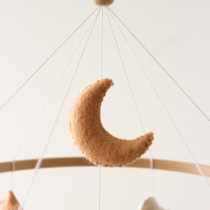 May include: A baby mobile featuring a crescent moon, star, and cloud made of soft, tan felt. The wooden hoop and bead add a natural touch. The mobile hangs from white strings, creating a whimsical nursery decoration.