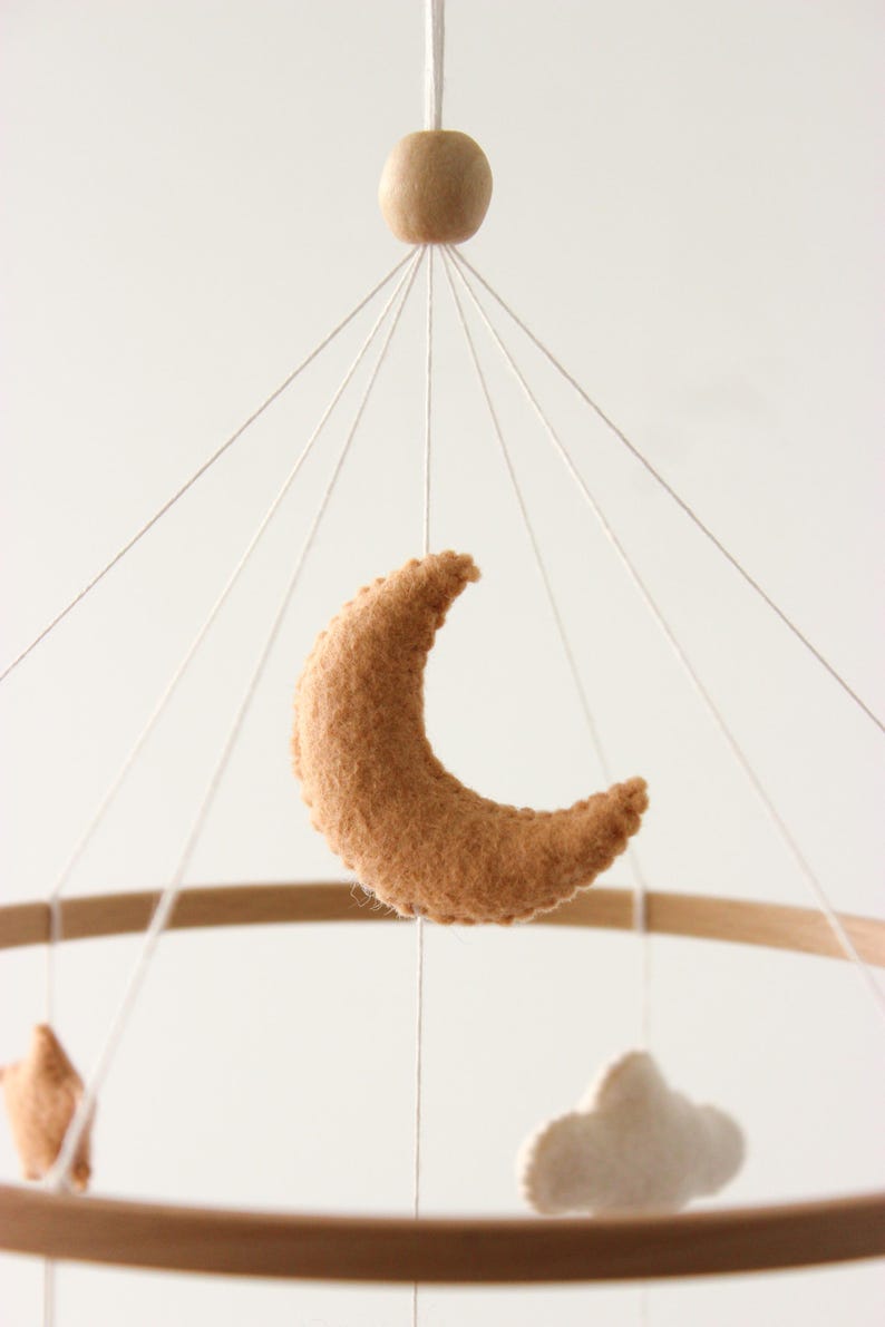 May include: A baby mobile featuring a crescent moon, star, and cloud made of soft, tan felt. The wooden hoop and bead add a natural touch. The mobile hangs from white strings, creating a whimsical nursery decoration.