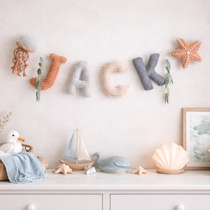 Puede incluir: Decoración de guardería con una guirnalda que deletrea "JACK" con letras de ganchillo y adornos marinos. La guirnalda incluye una medusa, una estrella de mar y vegetación. Debajo, una cómoda blanca exhibe una cesta con un pato, un velero y una figura de ballena.