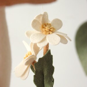 May include: A handmade felt flower mobile featuring two off-white flowers with yellow centers, a brown stem, and a green leaf. The mobile is suspended by a thin string, perfect for nursery decor.