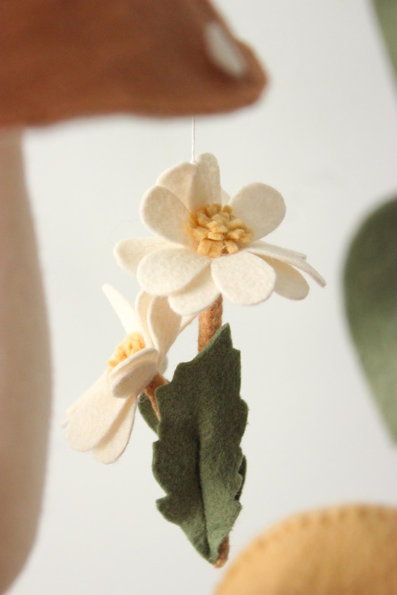 May include: A handmade felt flower mobile featuring two off-white flowers with yellow centers, a brown stem, and a green leaf. The mobile is suspended by a thin string, perfect for nursery decor.