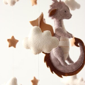 May include: A handmade baby mobile featuring a felt dragon, clouds, and stars in neutral tones. The dragon is gray with brown accents, and the stars and clouds are beige and white. The mobile is suspended from a wooden hoop.