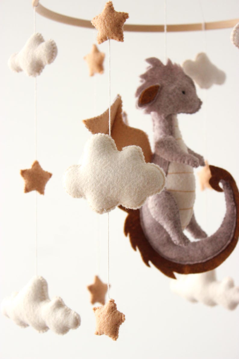 May include: A handmade baby mobile featuring a felt dragon, clouds, and stars in neutral tones. The dragon is gray with brown accents, and the stars and clouds are beige and white. The mobile is suspended from a wooden hoop.