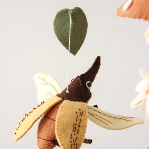 May include: A felt mobile featuring a brown and tan beetle with cream-colored wings, suspended by a white string. The mobile also includes green felt leaves and a mushroom.