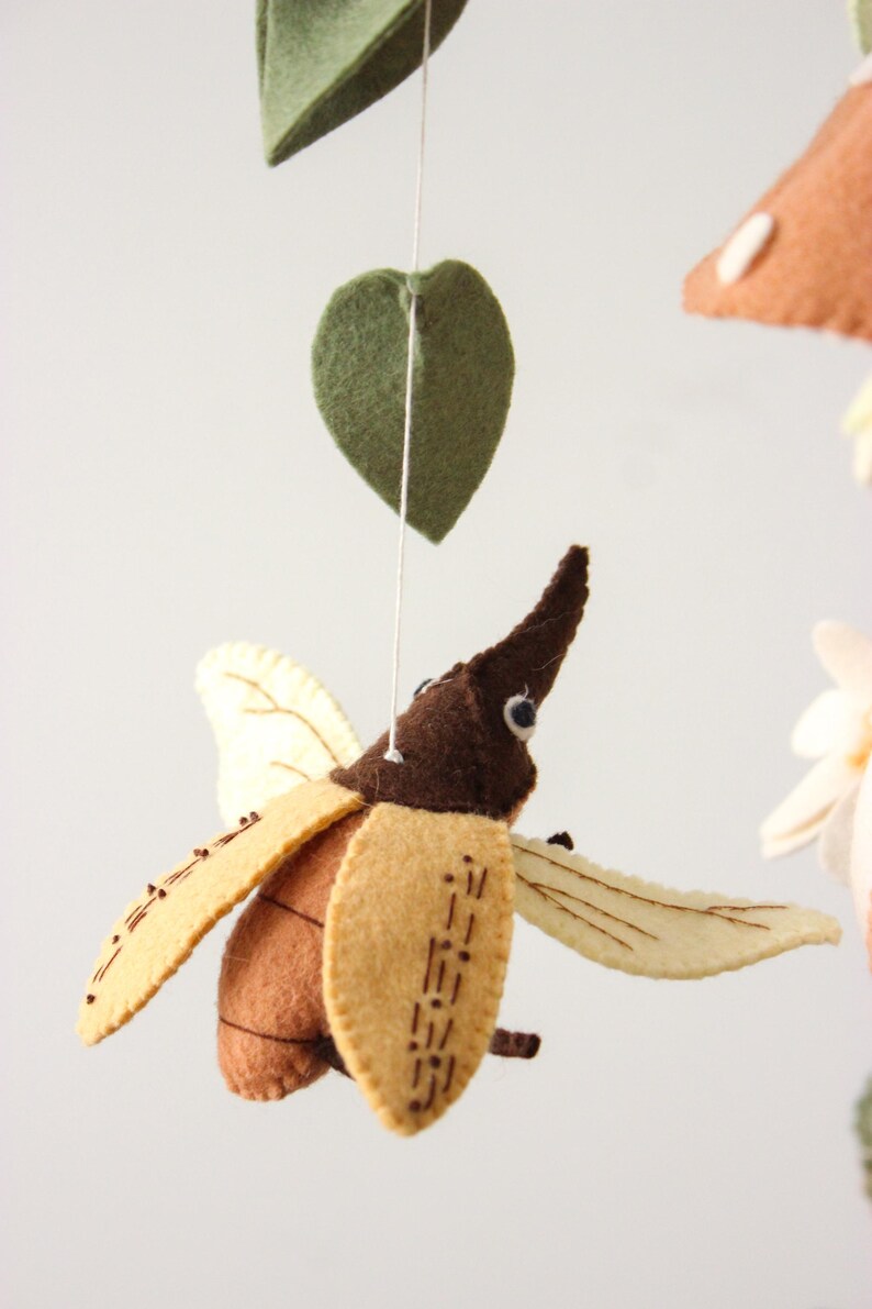 May include: A felt mobile featuring a brown and tan beetle with cream-colored wings, suspended by a white string. The mobile also includes green felt leaves and a mushroom.