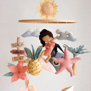 May include: A wooden mobile with a felt surfer girl, pineapple, starfish, seashell, and flower. The mobile also includes a felt sun and clouds. The signs on the mobile read "Beach", "Hawaii", and "Surf".