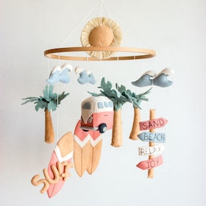 Op de afbeelding: Handgemaakte baby mobile met een strandthema. Het bevat een zon, wolken, palmbomen, surfplanken, een camperbusje en richtingborden met de tekst "Sand, Beach, Relax, Joy". De mobile is gemaakt van vilt.