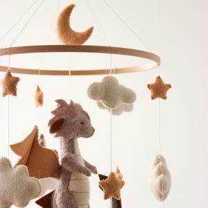 May include: A baby mobile featuring a felt dragon, clouds, stars, and a crescent moon. The dragon is gray with brown wings and tail. The mobile has a wooden ring and is suspended by strings.
