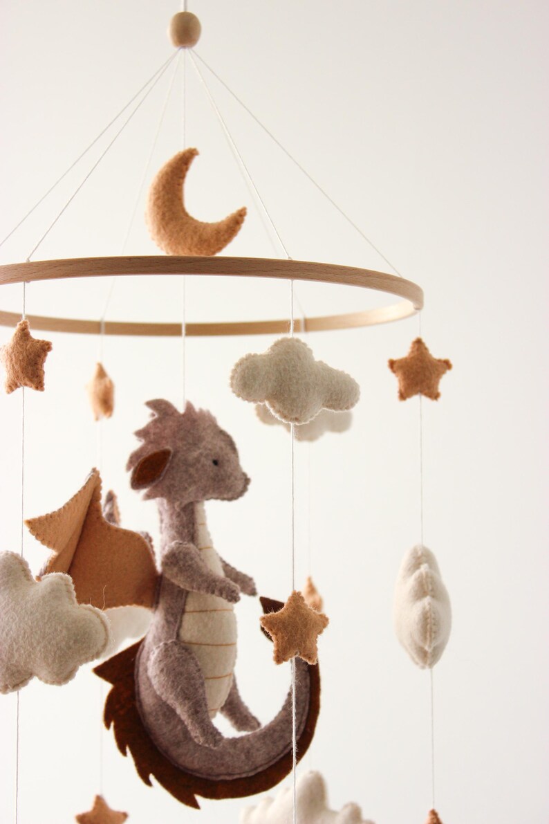 May include: A baby mobile featuring a felt dragon, clouds, stars, and a crescent moon. The dragon is gray with brown wings and tail. The mobile has a wooden ring and is suspended by strings.