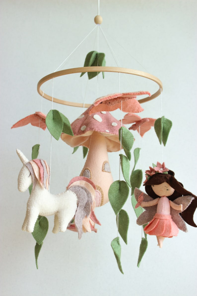 May include: A pink and green felt baby mobile featuring a mushroom, unicorn, and fairy. The mushroom has a white door and a pink and white polka dot top. The unicorn is white with a pink and purple mane and tail. The fairy has brown hair, a pink dress, and green wings.