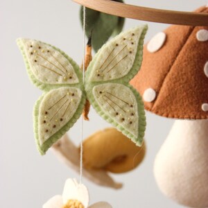 May include: A handmade baby mobile featuring felted elements. The mobile includes a light green butterfly with embroidered details, a brown mushroom with white spots, a white flower, and a yellow snail. The elements are suspended from a wooden hoop.