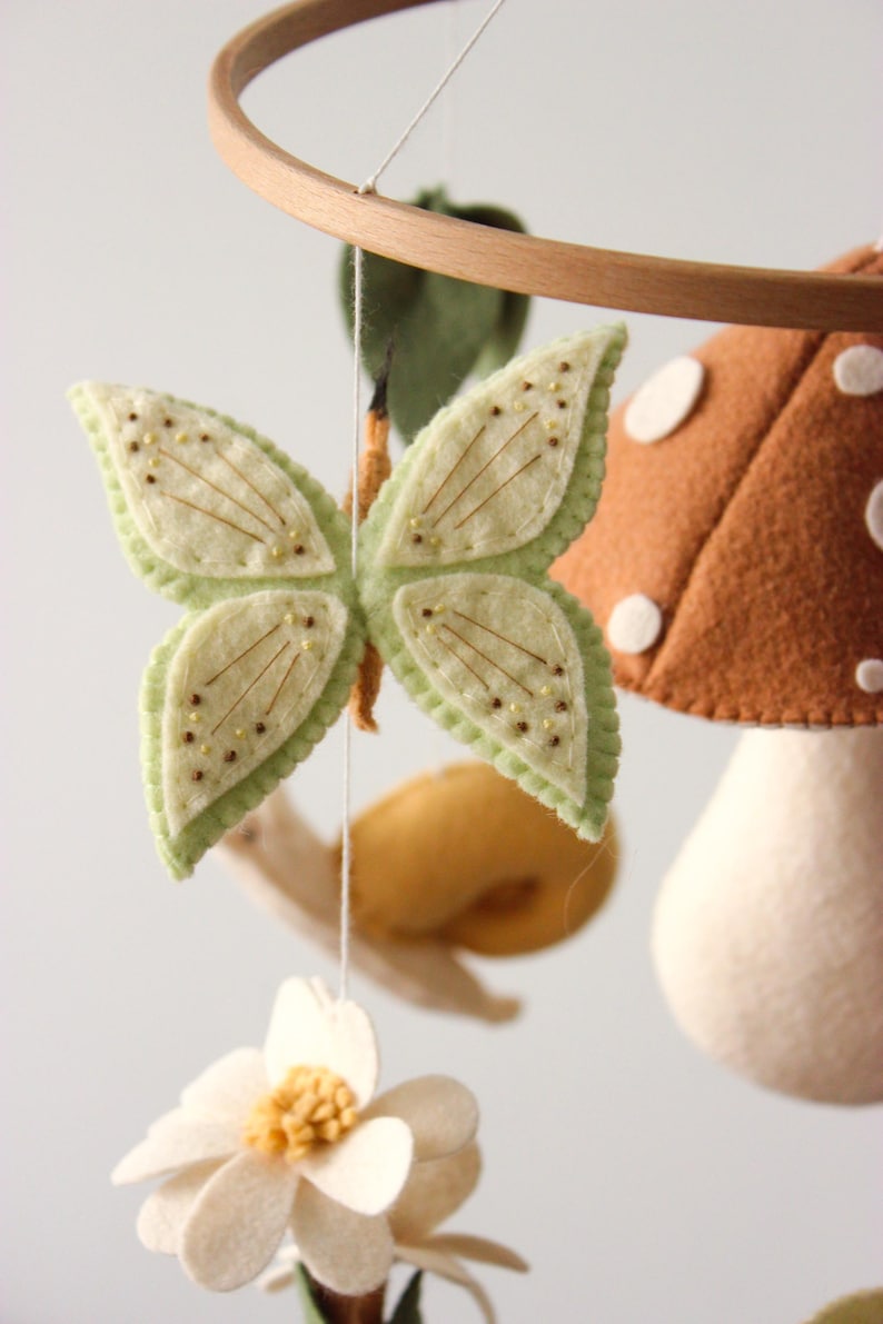 May include: A handmade baby mobile featuring felted elements. The mobile includes a light green butterfly with embroidered details, a brown mushroom with white spots, a white flower, and a yellow snail. The elements are suspended from a wooden hoop.