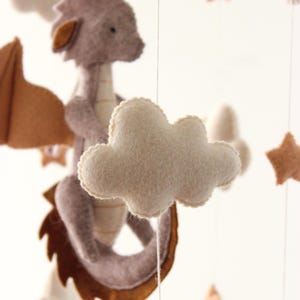 May include: A handmade baby mobile featuring a felt dragon with brown wings, a light purple body, and a cream-colored belly. The mobile also includes cream-colored clouds and tan stars, all suspended by strings.