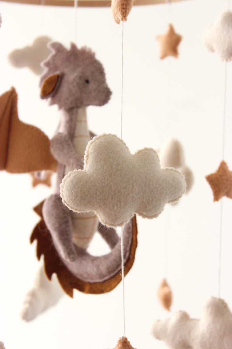 May include: A handmade baby mobile featuring a felt dragon with brown wings, a light purple body, and a cream-colored belly. The mobile also includes cream-colored clouds and tan stars, all suspended by strings.